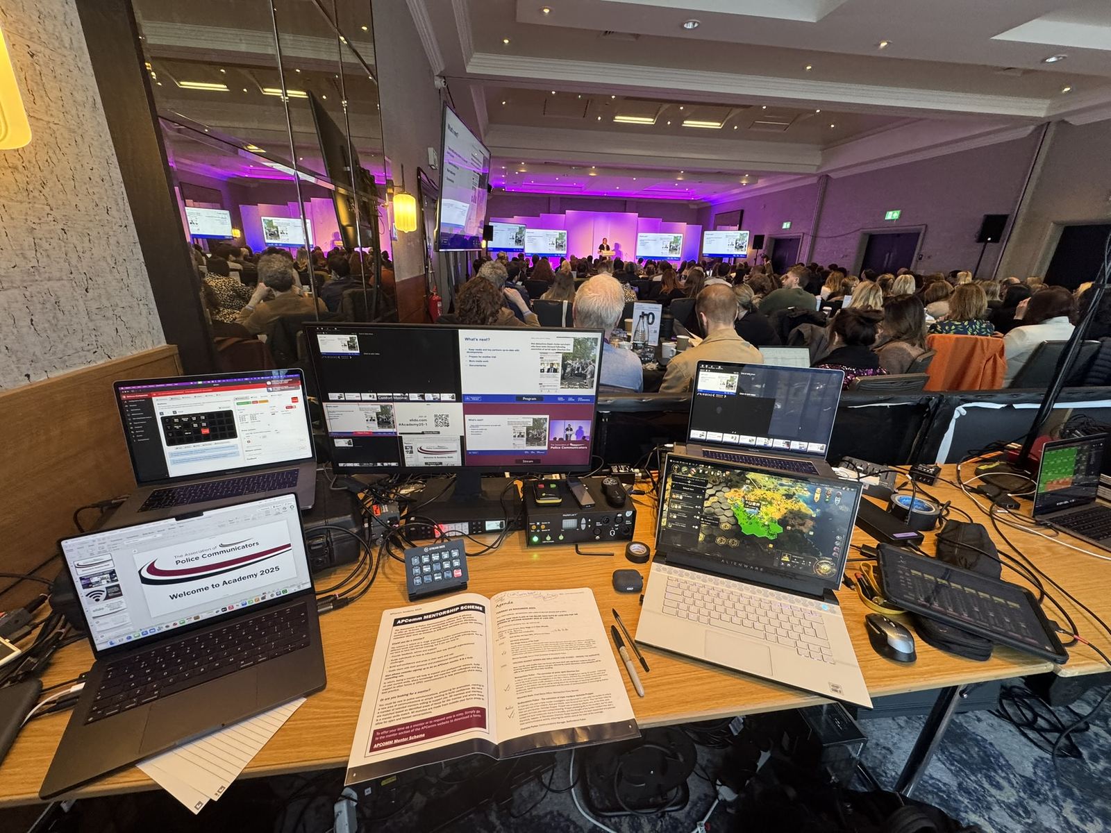 Multi-laptop live streaming and vision-mixing control desk at a conference, audience in the background