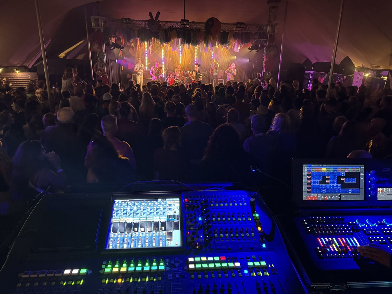Full band performing under festival tent lighting, digital mixer and lighting desk in foreground