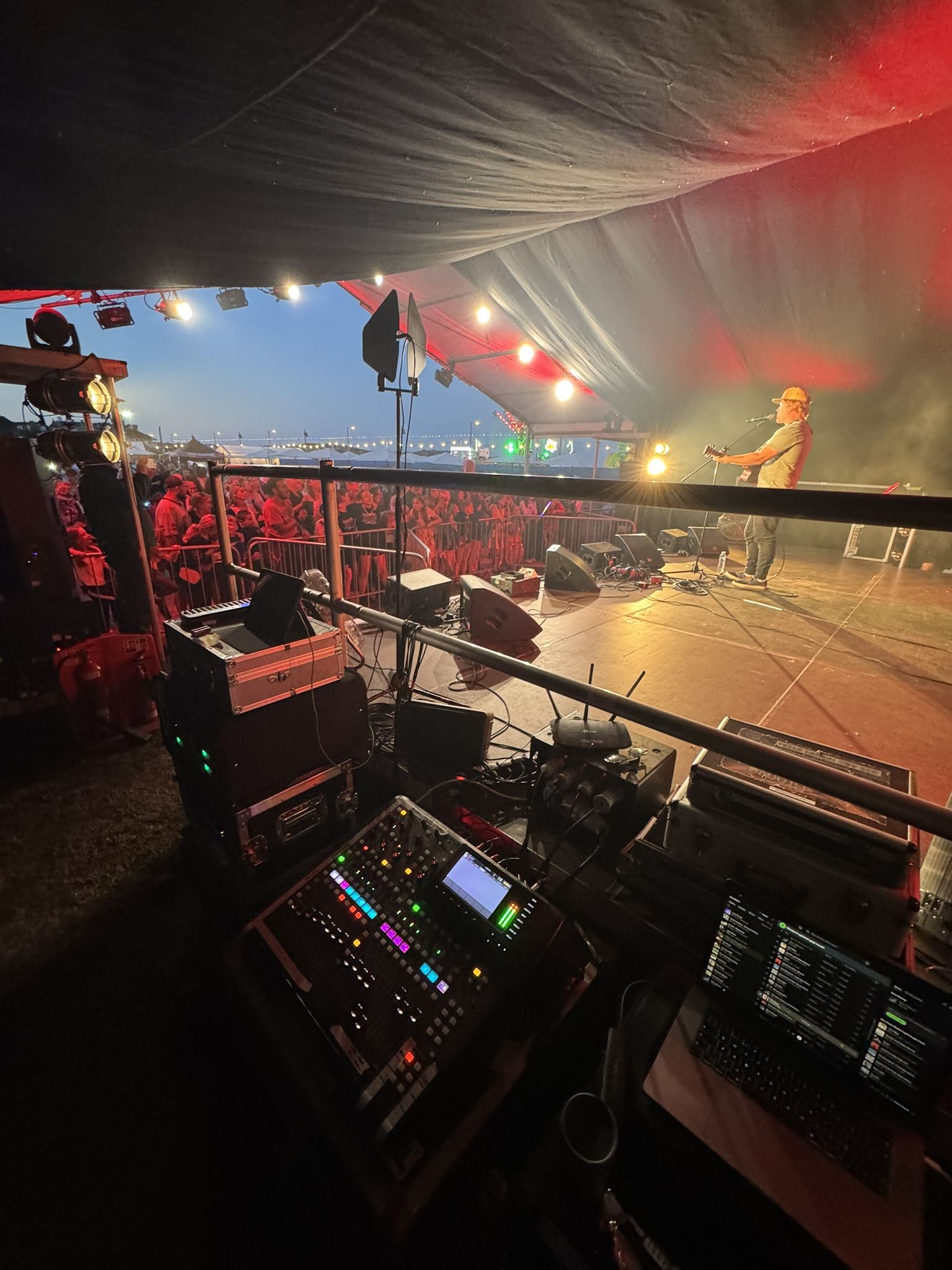 Solo musician performing on an outdoor festival stage at dusk, mixing desk in foreground